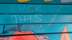 Inexpert graffiti on a blue park bench reads "Fuck this town"